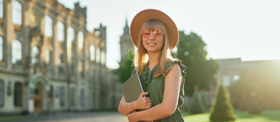 Blonde student with pink sunglasses & sunhat smiling next to ancient building