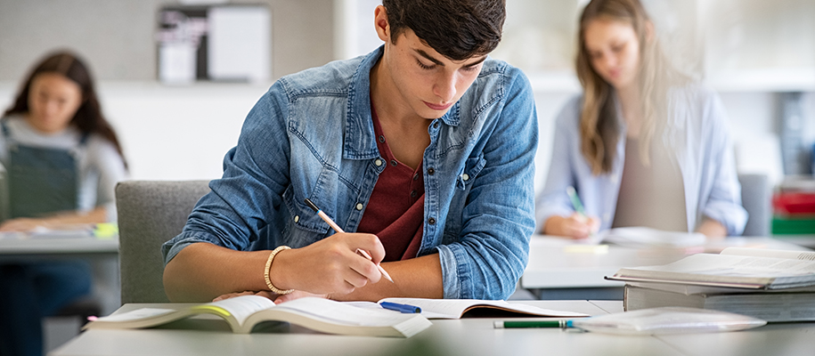 White male in denim shirt at desk in classroom with books and pens, studying