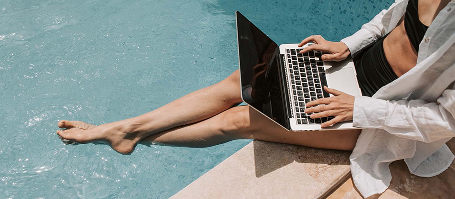 White woman in black bikini, white button-up shirt sitting by pool with laptop