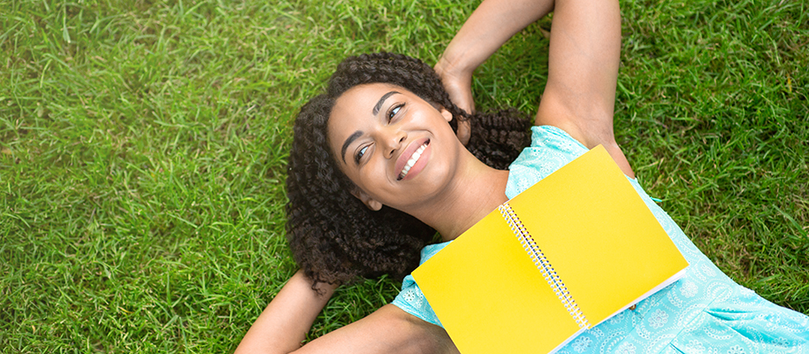 Black woman in blue shirt laying in green grass with yellow notebook on chest