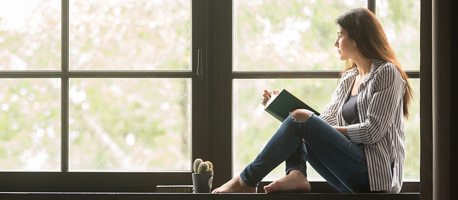 Brunette female with book sitting at window looking outside longingly