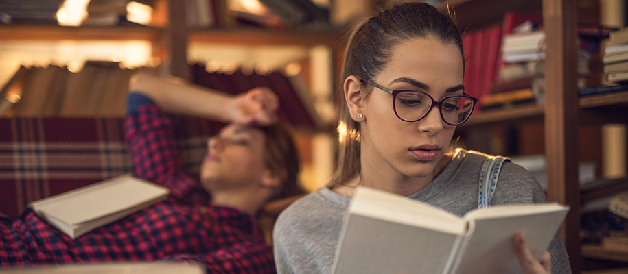 White woman in glasses reading book, friend asleep with book on couch behind
