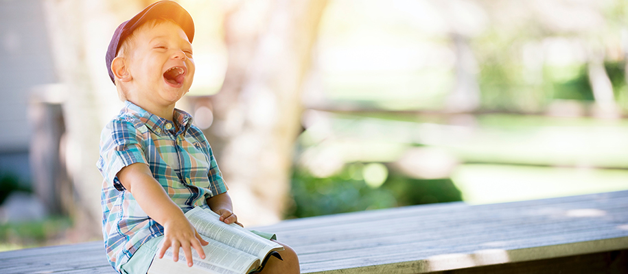 Little boy in baseball hat and green plaid shirt with open book smiling on a ben