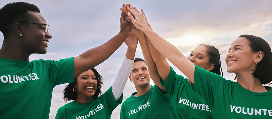 Five diverse young people in green volunteer shirts high fiving in a group