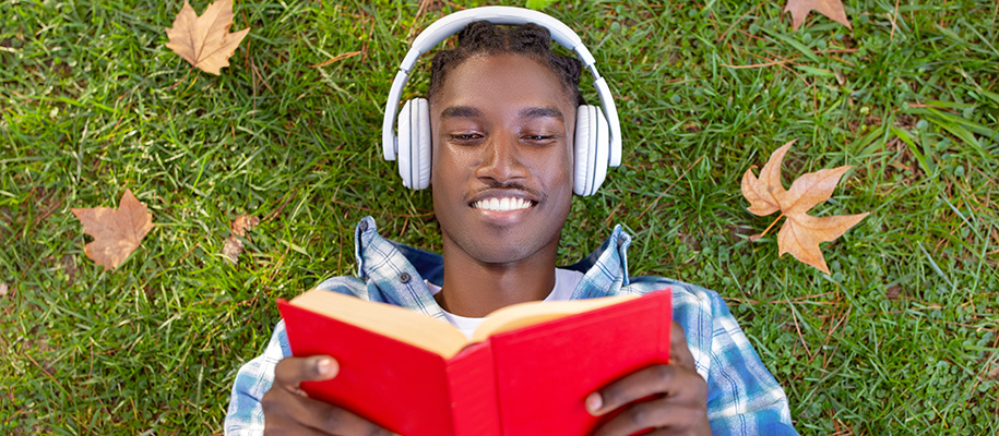 Black male in blue plaid shirt laying in grass with red book & white headphones