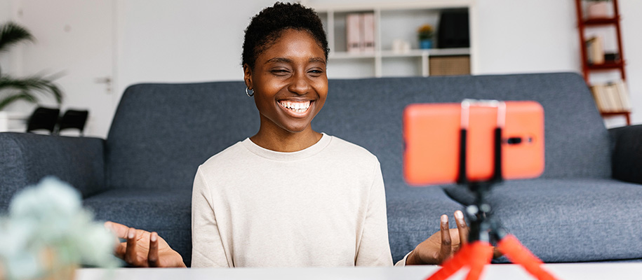 Black woman in white sweater smiling at red phone on tripod, filming video