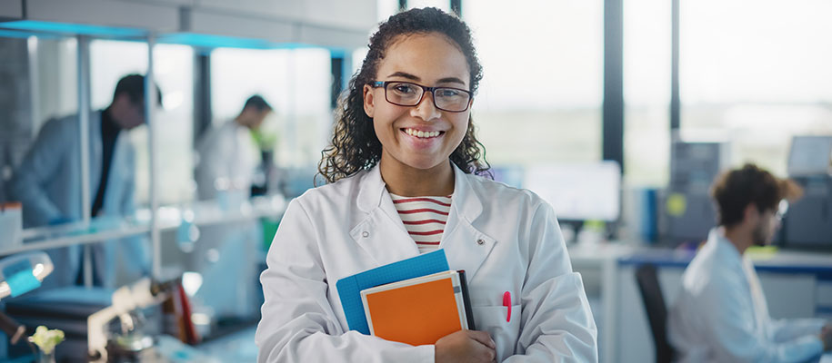 Black female scientist in white coat and glasses, smiling, holding books in lab
