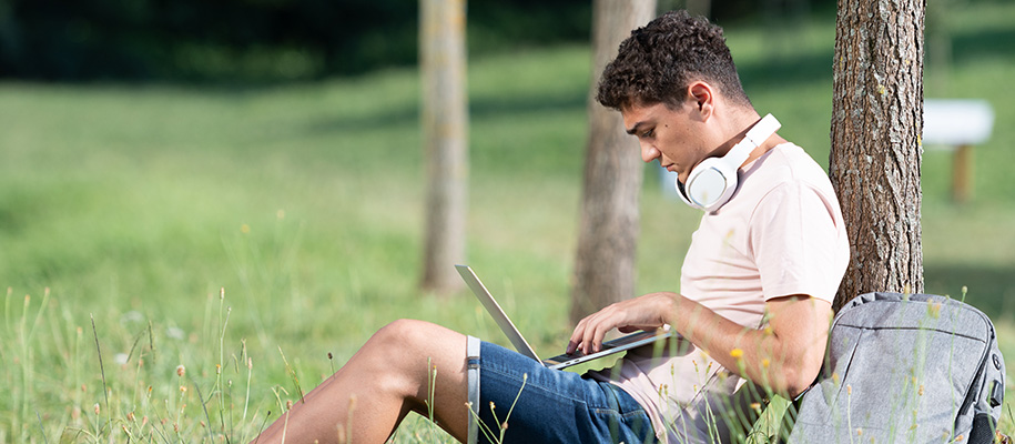 Hispanic high school boy typing on computer against tree in park with backpack