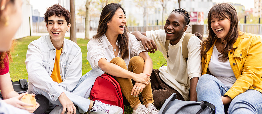 Six diverse students with bags on green college campus laughing together