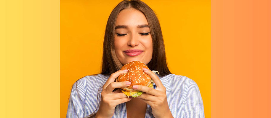 Brunette female holding cheeseburger with two hands, eyes closed, smiling