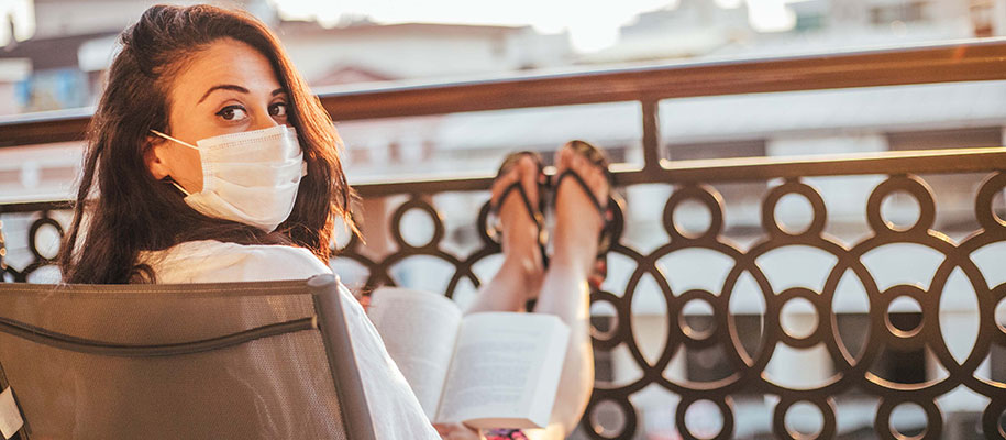 Woman wearing medical face mask turning head to camera, reading book on balcony