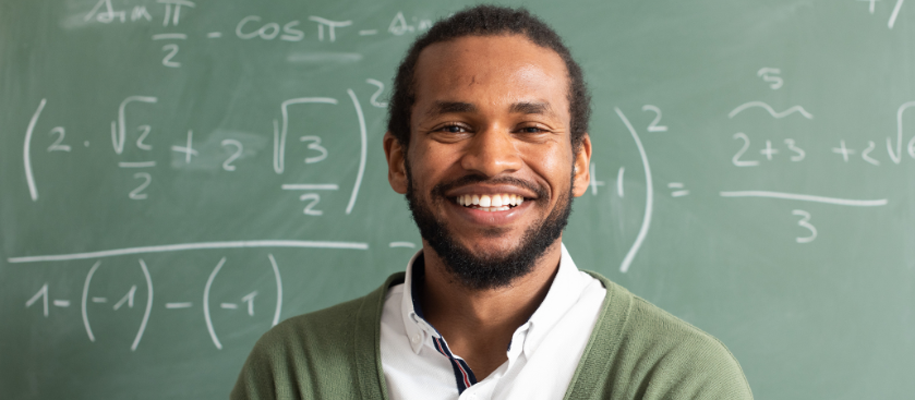 Black male teacher in sweater smiling in front of math formulas on chalkboard