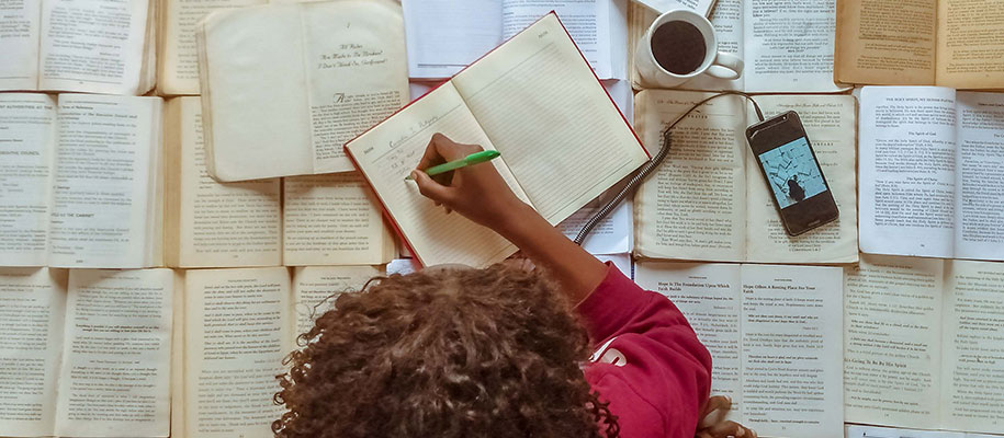 Black woman writing in book with many open books, coffee cup, and phone on desk