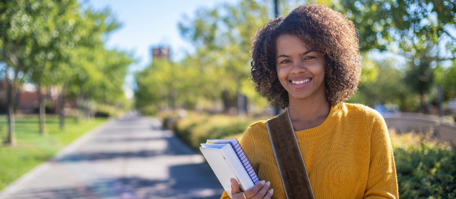 Black female college student with curly bob & yellow sweater on campus with book