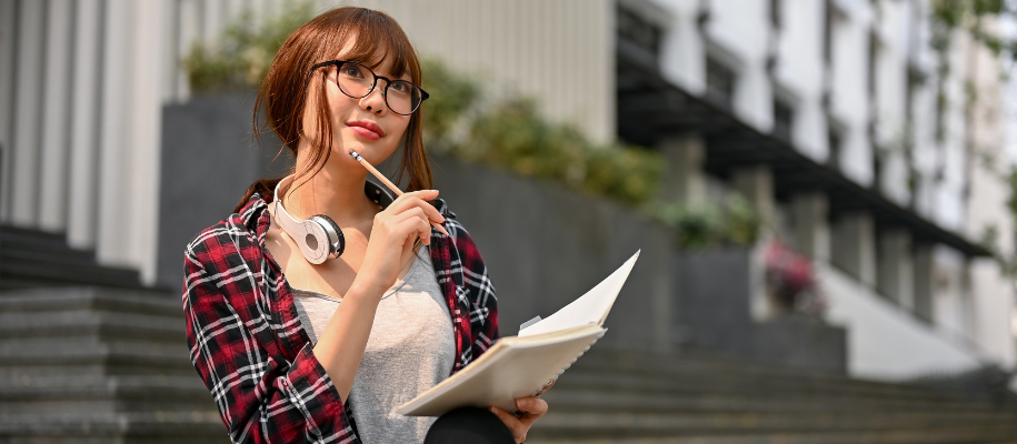 Asian female student with glasses outside, holding notebook with pencil to chin