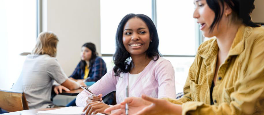 Black female taking notes, smiling at White female talking animatedly