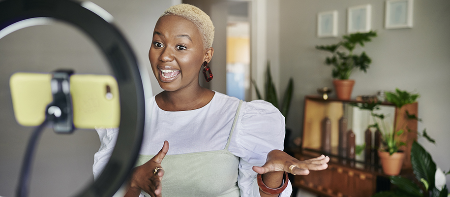 Black woman with shaved blonde hair in dress, filming with phone and ring light