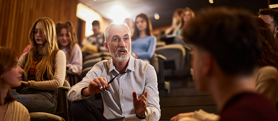 White male professor sits on lecture hall steps engaging with students in class