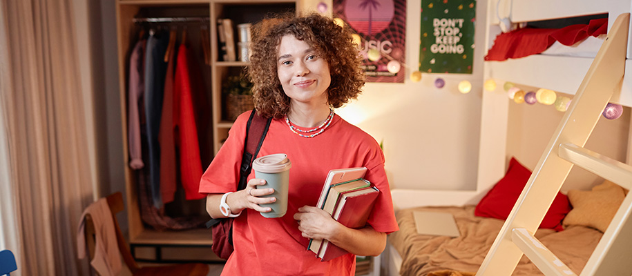 Woman with curly hair holding coffee, books in dorm room with bunk beds