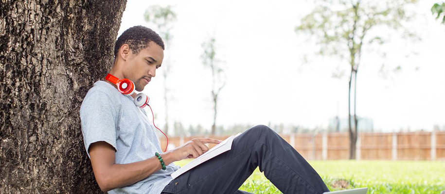 Black man with headphones around neck, reading textbook sitting against tree