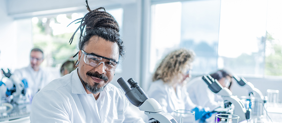 Black man with bun of dreadlocks in lab with microscopes and other scientists