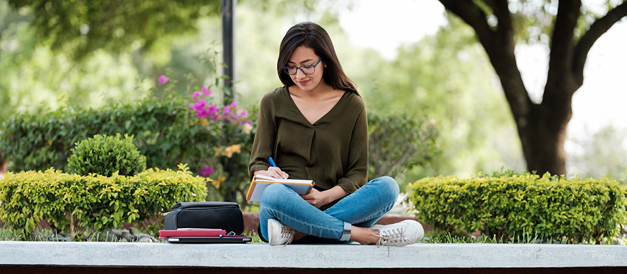 Hispanic woman with glasses, cross-legged on outdoor wall writing in notebook