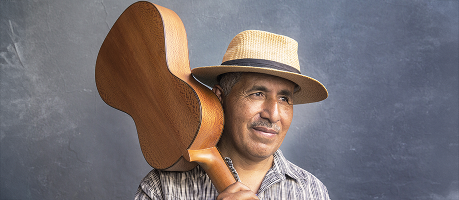 Latino man with straw hat, small guitar over shoulder, in a plaid shirt