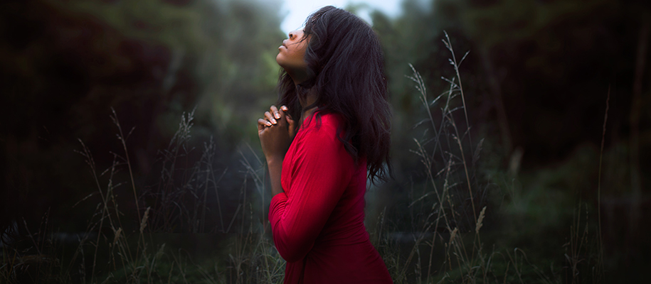 Black woman in red dress outside clasping hands in prayer, head tilted to sky