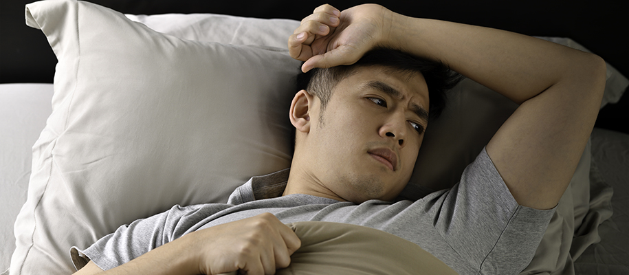 Young Asian man laying in bed with arm on forehead, looking concerned