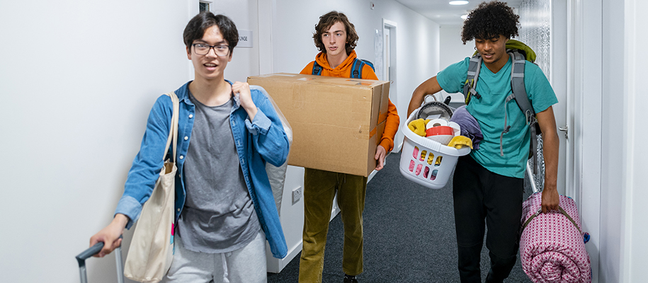 Asian, White & Black male students walking in dorm hallway with laundry and bags