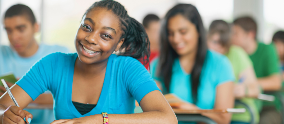 Black girl in blue shirt, smiling at camera in desk at foreground of classroom