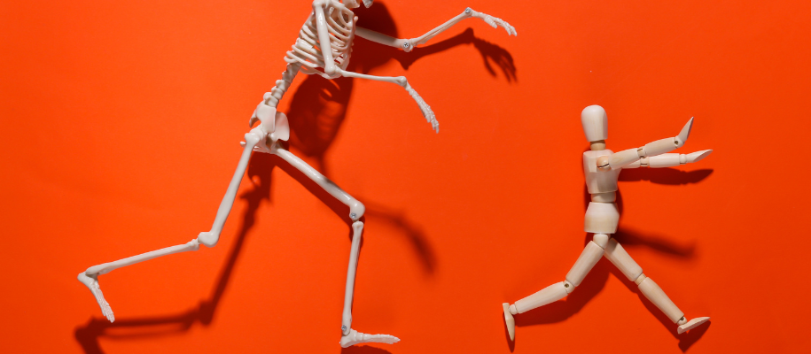 Plastic skeleton chasing posable wooden man on orange background with shadows