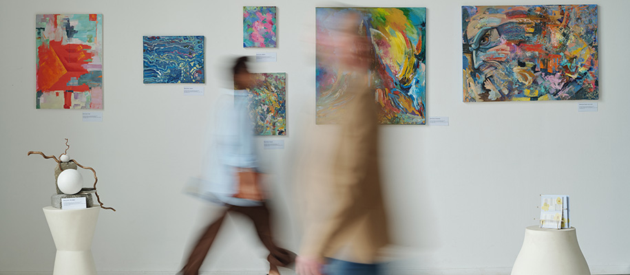 Two blurred people walking past art on wall of exhibit in white museum