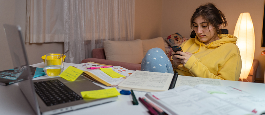 Multiracial woman in yellow sweatshirt, scrolling on phone at messy desk