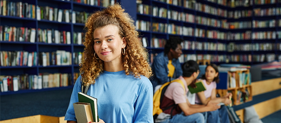 Curly-haired woman in blue shirt, holding books in library, students behind