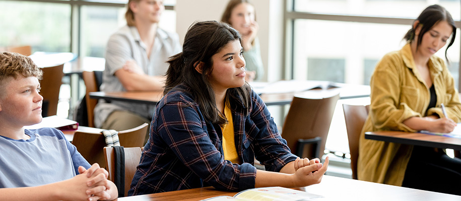 Focus on young Hispanic woman in plaid shirt in college classroom with students