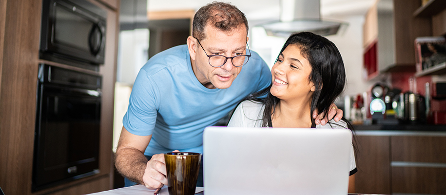 Mixed-race woman smiling at White dad with glasses in kitchen, looking at laptop