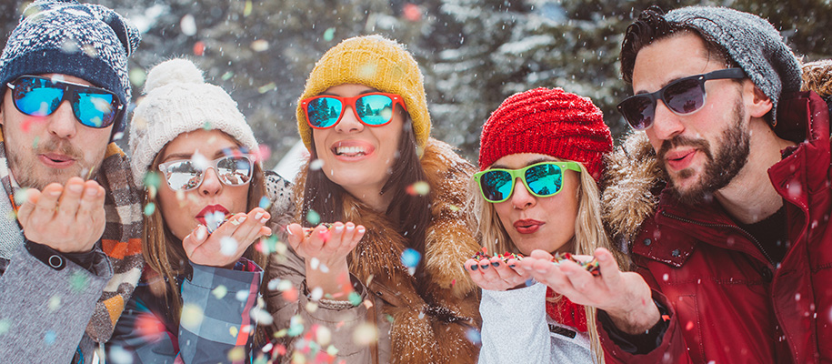 Five friends outside in winter in coats, hats, sunglasses, blowing confetti