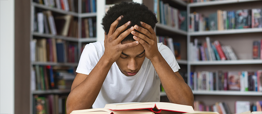 Young Black man in white T-shirt holds head in hands stressed in library