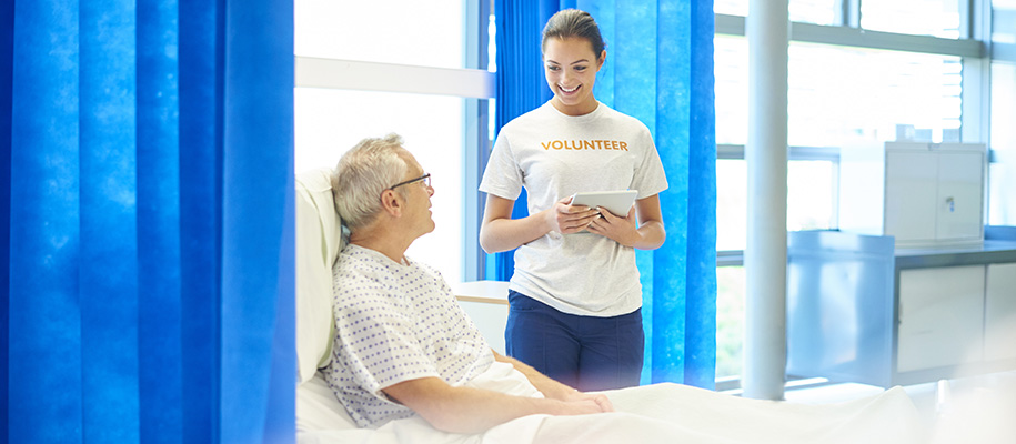 White woman in volunteer shirt with table talking with White man in hospital bed