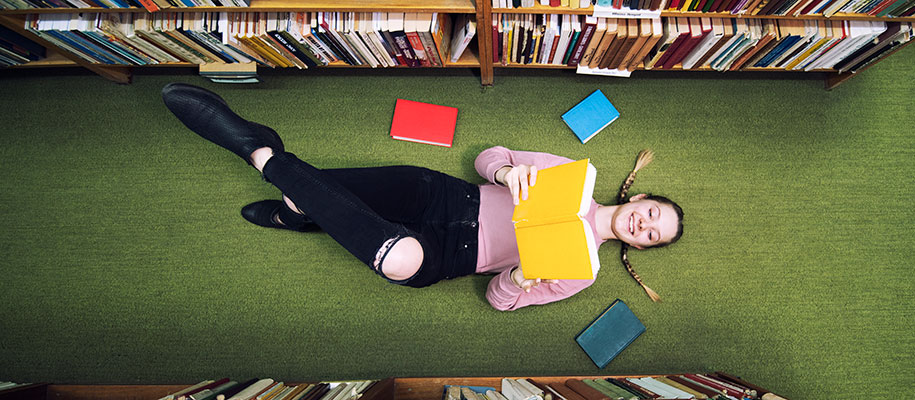 Overhead of blonde female student lying on library floor between shelves reading