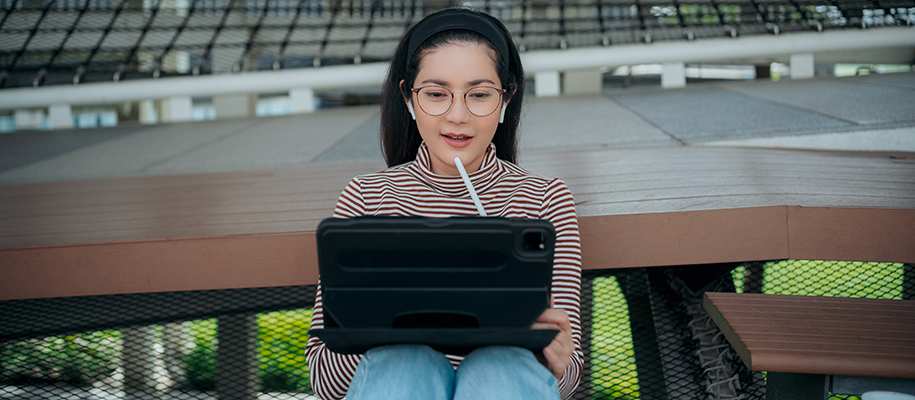 Asian woman in striped shirt and glasses in park with tablet, pencil to chin