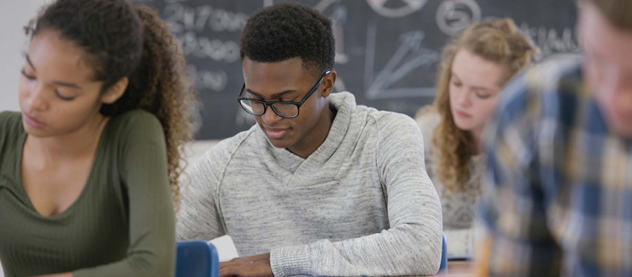 Black male in sweater and glasses sitting with peers in classroom taking test