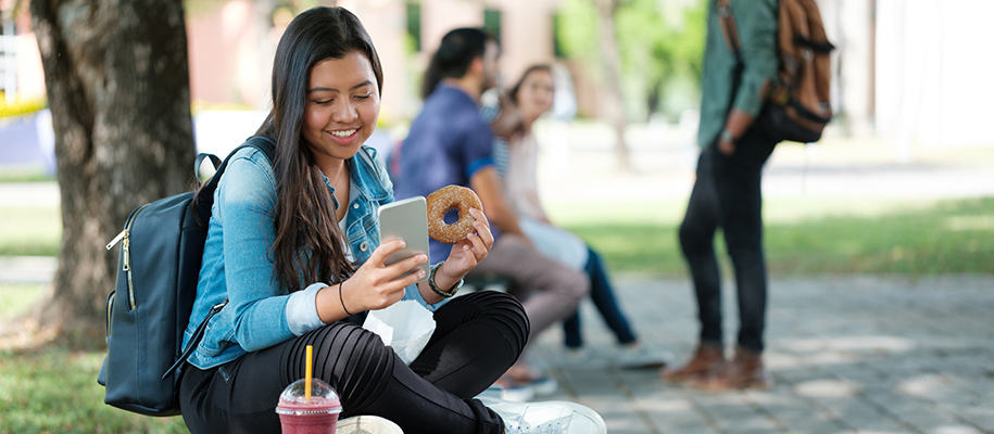 Latina woman on campus holding phone, donut, with a smoothie on bench
