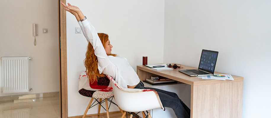 Red-headed woman in white shirt stretching with hands over head at desk in dorm