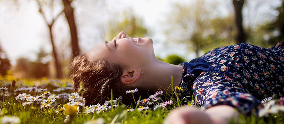 White female in flowered dress lying outside in daisy field smiling, eyes closed