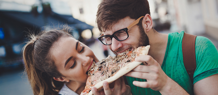Woman and man in green T-shirt and glasses both biting same slice of pizza