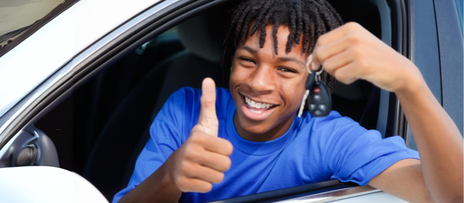 Young Black male teen in white car, holding keys through window and smiling
