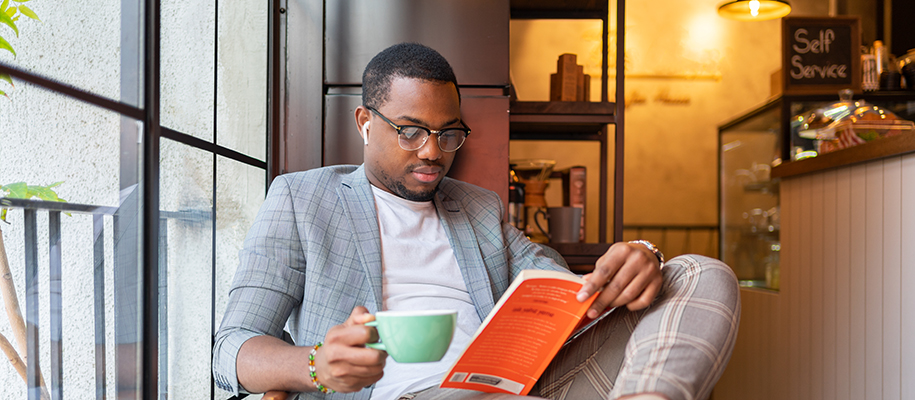 Black man in nice blazer with glasses reading book and drinking coffee in shop