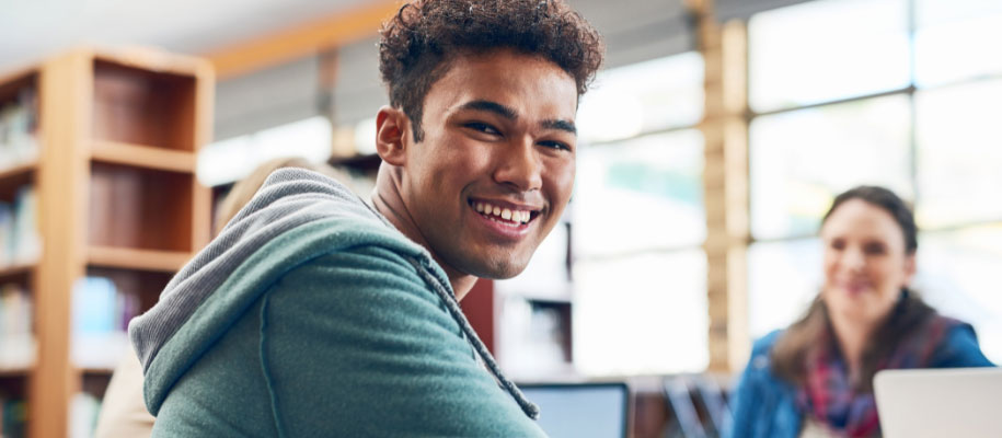 Male student of color sitting in library smiling at camera with other student